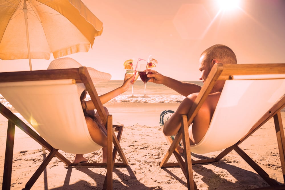 Happy couple on the beach clinking their glasses while relaxing on their deck chairs
